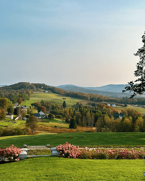 A scenic view of rolling hills and houses under a clear sky, framed by colorful autumn foliage.