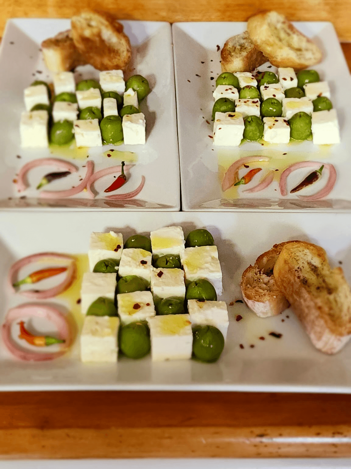 Three plates of feta cheese cubes, green olives, and pickled onions, garnished with herbs and served with toasted bread.