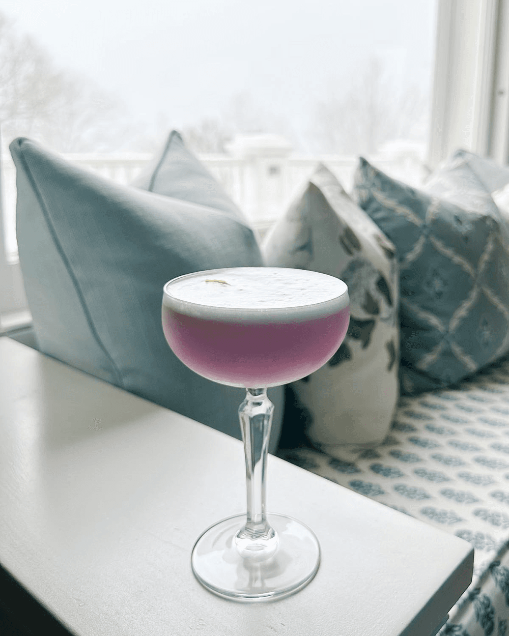 A pink cocktail with a frothy top sits on a table next to decorative cushions.