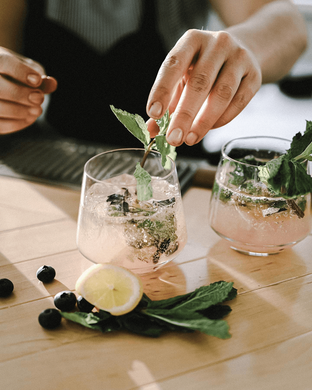 A hand garnishes a refreshing pink cocktail with mint leaves, alongside fresh berries and lemon on a wooden table.