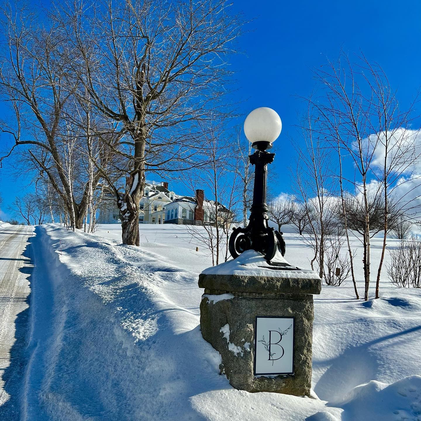 A lamp post stands beside a snowy path leading to a large house under a clear blue sky.