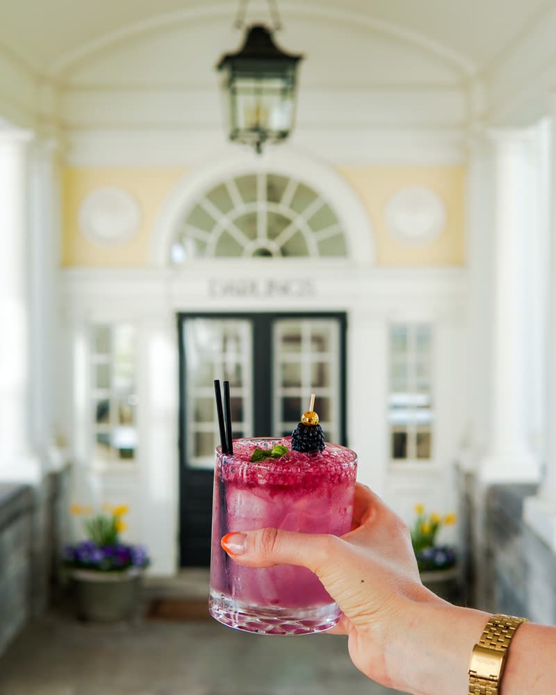 A hand holds a vibrant pink cocktail with blackberries in front of a bright doorway.