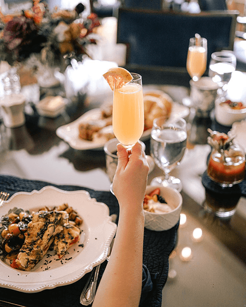 A hand holding a glass of mimosa over a beautifully set dining table with various dishes.