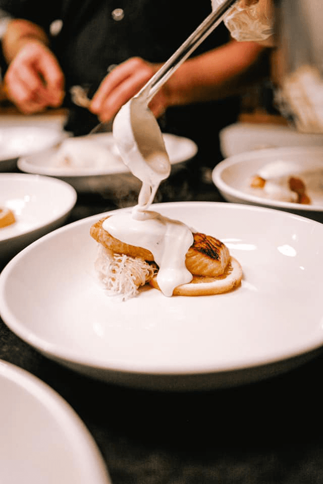 A chef drizzles sauce over a plated dish featuring seared fish and garnishes.
