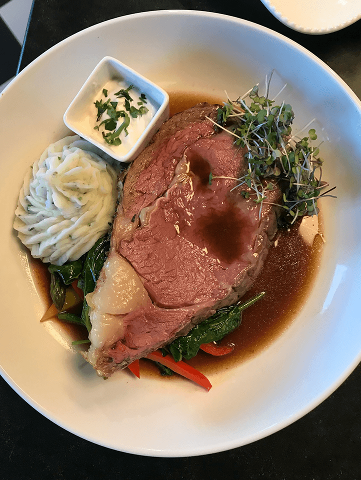 A plated dish featuring prime rib, mashed potatoes, sautéed vegetables, and a small bowl of sauce.