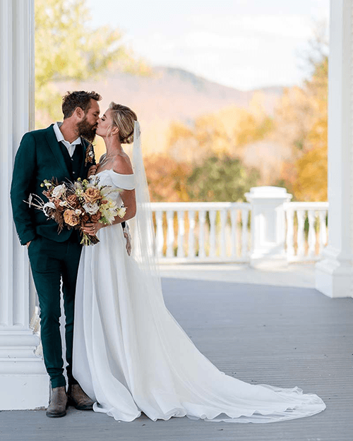 A newlywed couple kisses while holding a bouquet against a backdrop of autumn foliage.